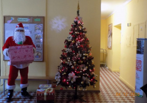 Mikołaj stoi przy choince i trzyma prezent.