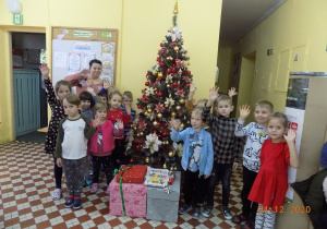 Dzieci z nauczycielką z grupy skrzatów stoją na tle choinki i prezentów i machają do obiektywu.