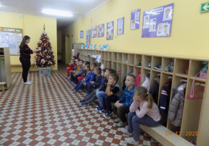 Dzieci z grupy elfów siedzą na ławce w holu przedszkolnym- nauczycielka czyta list od Świętego Mikołaja.