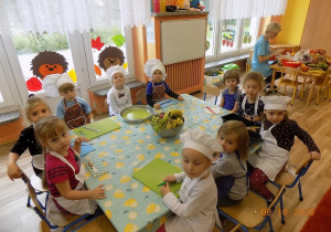 Dzieci siedzą przy stole w czapkach i fartuszkach kucharskich.