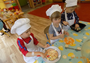 Dzieci w czapkach i fartuszkach kucharskich kroją owoce.