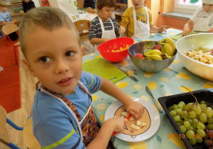 Dzieci w czapkach i fartuszkach kucharskich kroją owoce, chłopiec spogląda w obiektyw.