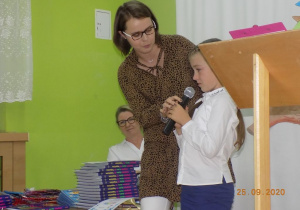 Nauczycielka podtrzymuje mikrofon dzewczynce mówiącej wiersz.