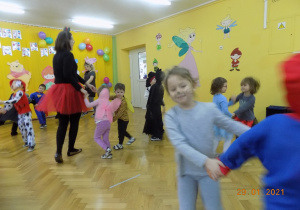 Dzieci z nauczycielkami w strojach karnawałowych tańczą na sali gimnstaycznej w małych kółeczkach.