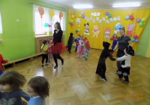 Dzieci z nauczycielkami w strojach karnawałowych tańczą na sali gimnstaycznej w małych kółeczkach.