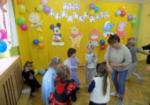 Dzieci w grupy skrzatów i nauczycielka tańczą na balu karnawałowym.