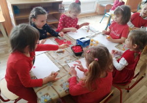 Dzieci siedzą przy stołach i naklejają kawałki czerwonej bibuły na kontur serca.