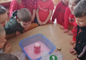 Dzieci ustawione wokół jednego stołu wykonują eksperyment z czerwonym barwnikiem- wulkan.