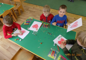 Trzech chłopców i jedna dziewczynka z grupy skrzatów siedzą przy stole i kolorują serce.