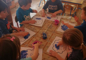 Dzieci siedzą przy stołach i malują paluszkiem zamoczonym w niebieskiej farbie.