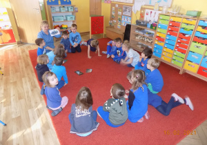 Dzieci siedzą w kole na dywanie, nauczycielka pokazuje niebieską ilustrację.