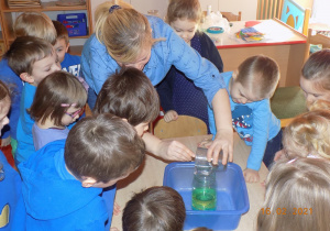 Dzieci ustawione wokół jednego stołu, nauczycielka przelewa płyn ze szklanki do słoika.