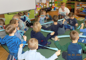 Dzieci z grupy skrzatów bawią się niebieskimi paskami bibuły na dywanie w rytm muzyki.