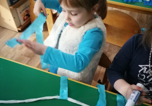 Dziewczynka przykleja niebieskie paski bibuły i przykleja do sznurka.