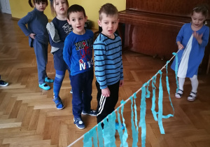 Dzieci stoją w rzędzie, a dwie dziewczynki trzymają sznurek z niebieskimi paskami bibuły. Dzieci będą przebiegały pod paskami.