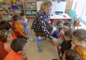 Dzieci z nauczycielką ustawione wokół stołu, jeden chłopiec zagląda do słoika.