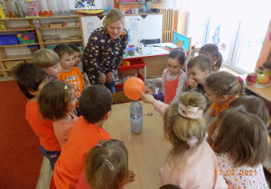 Dzieci z nauczycielką ustawione wokół stołu, obserwują jak rośnie balon założony na butelkę.