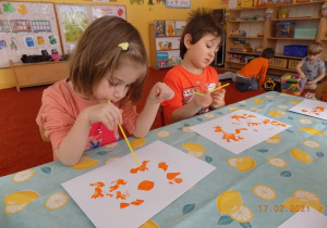 Dwoje dzieci siedzi przy stole i za pomocą słomki do napojów rozdmuchują kleksy z pomarańczowej farby.