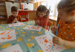 Troje dzieci siedzi przy stole i za pomocą słomki do napojów rozdmuchują kleksy z pomarańczowej farby.