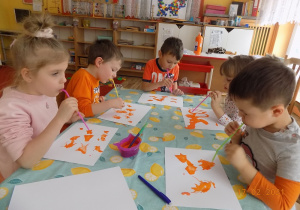Dzieci siedzą przy stole i za pomocą słomki do napojów rozdmuchują kleksy z pomarańczowej farby.