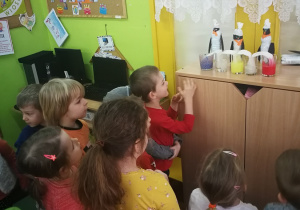 Dzieci z grupy trzeciej stoją i obserwują eksperyment z mieszaniem się kolorów w szklankach za przyczyną nasiąkania papieru.