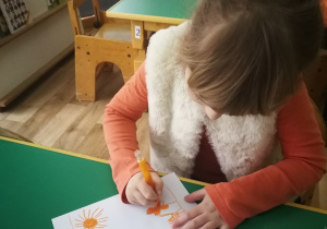 Dziewczynka z grupy trzeciej siedzi przy stole i maluje pomarańczowy obrazek