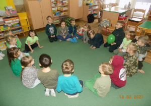 Dzieci z grupy trzeciej, ubrane na zielono siedzą w kole i grają w zielonego balona.