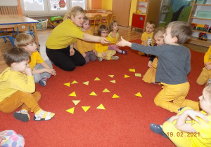 Dzieci siedzą w kole, na środku leżą żółte trójkąty, nauczycielka podaje dziecku jeden trójkąt.