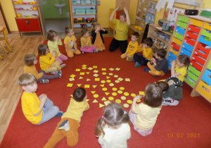 Dzieci z nauczycielką siedzą w kole, na środku leżą żółe figury- koła, trójkąty i kwadraty.