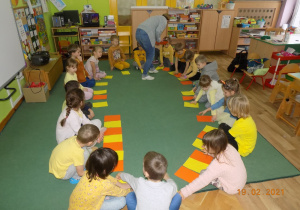 Dzieci z grupy trzeciej siedzą w kole, a nauczycielka rozkłada na podłodze kartki w raz w żółtym kolorze, a raz w pomarańczowym.