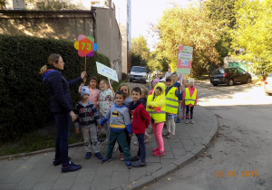 Dzieci spacerują z nauczycielką po osiedlowych ścieżkach. Nauczycielka trzyma w ręku transparent z numerem przedszkola 73. Dwójka dzieci trzyma transparenty, na których są hasła dotyczące ochrony środowiska