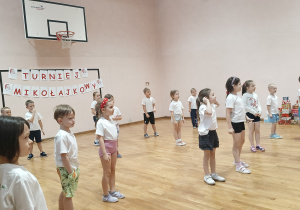 Dzieci stoją na sali gimnastycznej i wykonują sportową rozgrzewkę.