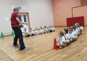 Dzieci siedzą w dwóch rzędach i czekają na kolejną konkurencję. Nauczycielka tłumaczy zadanie.