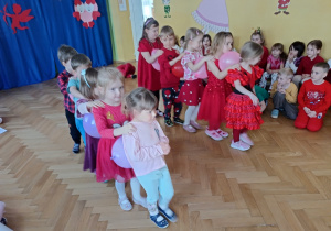 Dzieci w dwóch drużynach po pięcioro, ustawione jeden za drugi ścigają się z balonami ściśniętymi między sobą.