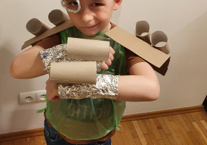 Chłopiec ma na głowie kask wykonany z folii metalowej. Niektóre elementy stroju wykonane są z rolek po ręczniku papierowym