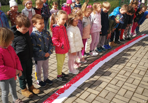 Dzieci z grupy drugiej stoją w szeregu przed flagami, które lażą na chodniku ułożone w dywan z flag