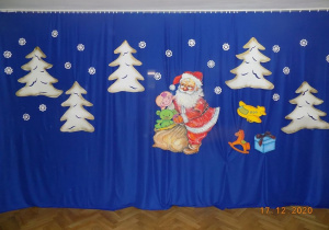 Dekoracja Bożonarodzeniowa. Na płótnie zawieszone są białe choinki, Mikołaj z workiem oraz zabawki z papieru.