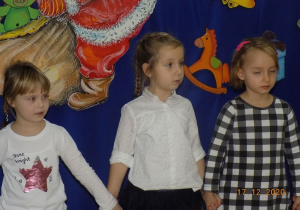 Trzy dziewczynki trzymają się za ręce i śpiewają Świąteczną piosenkę.