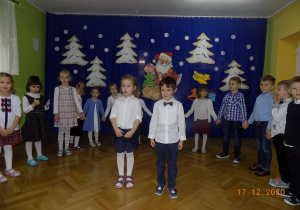Chłopiec i dziewczynka stoją z przodu i mówią wiersz Świąteczny, a pozostałe dzieci stoją za nimi i trzymają się za ręce.
