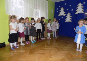 Dzieci przygotowują się do instrumentacji utworu muzycznego. Są ustawione na pozycjach. Dziewczynki stoją po lewej stronie.Na zdjęciu widać jedną dziewczynkę stojącą na środku z drewnianymi łyżkami