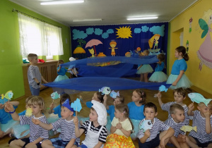 Dzieci siedzące z przodu poruszają papierowymi rybkami w rytm muzyki. Dzieci za nimi w parach tańczą z chustami.